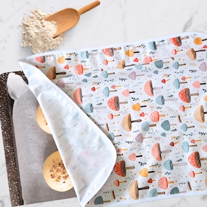 May include: A white mushroom-patterned baking mat partially covering a baking sheet with two round baked goods. A wooden scoop and a pile of flour are in the upper left corner. The mat features colorful mushroom illustrations.