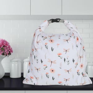 May include: White tote bag with a floral and dragonfly pattern. The bag features a black buckle closure and a gray handle. The design includes peach-colored flowers, green leaves, and dragonfly illustrations. The bag is sitting on a black surface.
