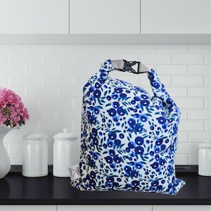 May include: A reusable lunch bag with a white background and a blue floral pattern. The bag has a gray buckle closure and a handle. The bag is sitting on a black surface.