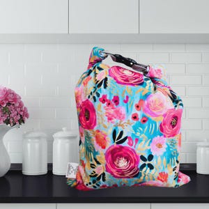 May include: A colorful floral drawstring bag with a black handle. The bag features a vibrant pattern of pink, red, and orange flowers on a turquoise background. The bag is made of fabric and is likely used for storage or carrying items.