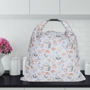 May include: A white tote bag with a repeating pattern of mushrooms in shades of orange, gray, and brown. The bag has a black handle and is sitting on a black surface. The background is a white brick wall.