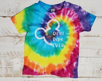 Peut inclure: Un t-shirt tie-dye avec un motif en spirale arc-en-ciel. Le t-shirt présente un contour blanc de la tête de Mickey Mouse avec le texte "BEST DAY EVER" à l'intérieur des oreilles.