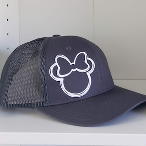 May include: A dark gray trucker hat with a white outline of a cartoon mouse head with a bow. The hat has a mesh back.