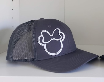 Peut inclure: Un chapeau de camionneur gris foncé avec un contour blanc d'une tête de souris de dessin animé avec un nœud. Le chapeau a un dos en maille.