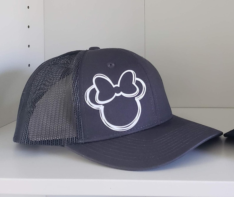 May include: A dark gray trucker hat with a white outline of a cartoon mouse head with a bow. The hat has a mesh back.