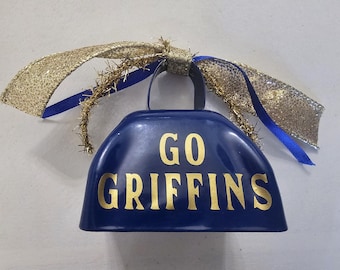 Peut inclure: Une cloche de vache bleue avec un nœud et un ruban dorés à paillettes. La cloche de vache porte l'inscription "Go Griffins" en lettres dorées.