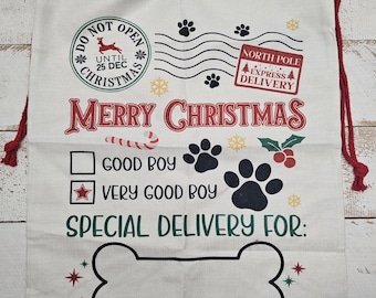 Peut inclure: Sac cadeau de Noël en toile blanche avec un cordon de serrage rouge. Le sac a un motif d'empreintes de pattes et le texte "Ne pas ouvrir avant le 25 décembre Noël", "Livraison express du pôle Nord", "Joyeux Noël", "Bon garçon", "Très bon garçon", et "Livraison spéciale pour :" avec une forme d'os en bas.