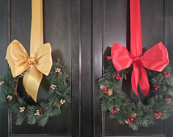 Peut inclure: Deux couronnes de Noël avec des branches de pin vertes et des nœuds rouges et dorés accrochés à une porte en bois foncé.