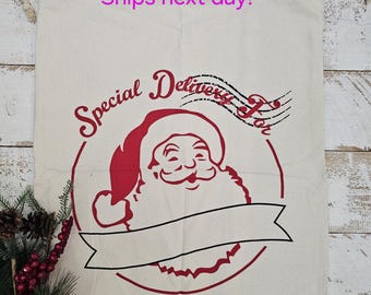 Peut inclure: Un sac de Noël en toile blanche avec des bordures rouges et une illustration rouge et blanche du visage du Père Noël. Le sac porte l'inscription "Special Delivery For" et "IF UNDELIVERED PLEASE RETURN TO THE North Pole".