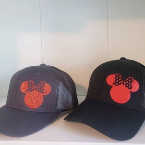 May include: Two black mesh trucker hats with red and black Minnie Mouse silhouettes. The Minnie Mouse silhouettes are made of glitter.