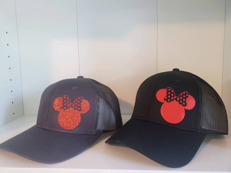 May include: Two black mesh trucker hats with red and black Minnie Mouse silhouettes. The Minnie Mouse silhouettes are made of glitter.