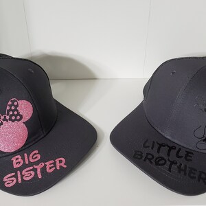 May include: Two black and gray trucker hats with mesh back panels. One hat features a pink Minnie Mouse silhouette with polka dots and the text "BIG SISTER". The other hat features a black Mickey Mouse silhouette in a dabbing pose and the text "LITTLE BROTHER".
