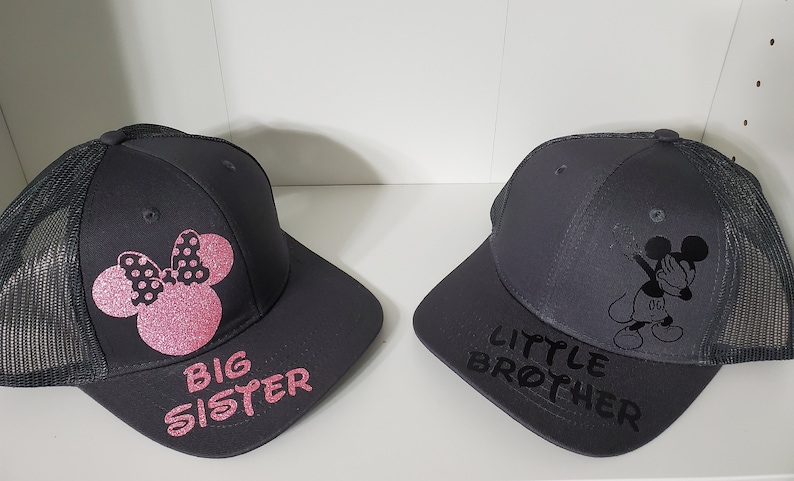 May include: Two black and gray trucker hats with mesh back panels. One hat features a pink Minnie Mouse silhouette with polka dots and the text "BIG SISTER". The other hat features a black Mickey Mouse silhouette in a dabbing pose and the text "LITTLE BROTHER".