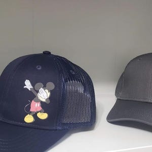 May include: Two black and gray baseball caps with a mesh back. Both caps feature a cartoon image of Mickey Mouse in a red and yellow outfit, doing the "dab" dance pose.