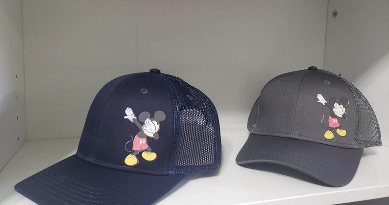May include: Two black and gray baseball caps with a mesh back. Both caps feature a cartoon image of Mickey Mouse in a red and yellow outfit, doing the "dab" dance pose.
