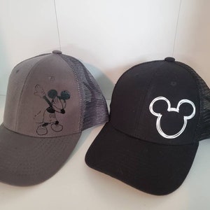 May include: Two black and gray baseball caps with a Mickey Mouse design. One cap features a black Mickey Mouse doing the dab, while the other cap features a white outline of Mickey Mouse's head.