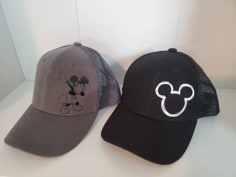 May include: Two black and gray baseball caps with a Mickey Mouse design. One cap features a black Mickey Mouse doing the dab, while the other cap features a white outline of Mickey Mouse's head.