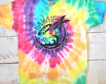Peut inclure: Un t-shirt tie-dye avec un motif noir et blanc d'un palmier, d'une planche de surf et de vagues. Le motif dit "Hawaii Aloha Family Vacation".