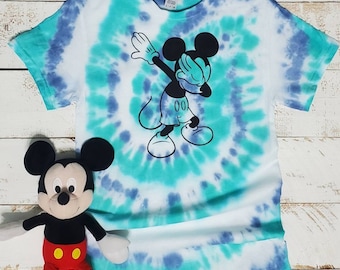 Peut inclure: Un t-shirt tie-dye avec une image en noir et blanc de Mickey Mouse faisant le "Dab". Le t-shirt est de couleur bleu clair et turquoise avec des tourbillons blancs.