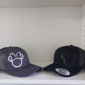 May include: Two black and gray trucker hats with embroidered designs. One hat features a white outline of Minnie Mouse's head with a bow. The other hat features a black outline of Mickey Mouse's head. Both hats have a black mesh back.