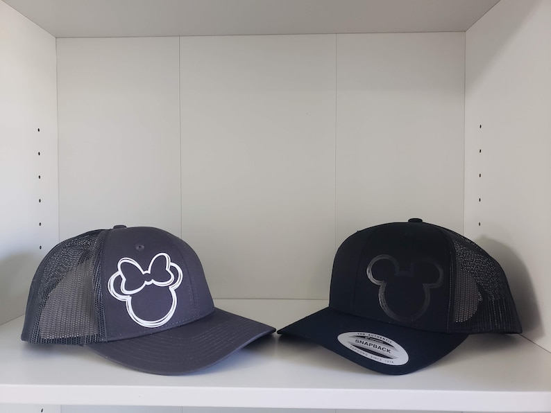 May include: Two black and gray trucker hats with embroidered designs. One hat features a white outline of Minnie Mouse's head with a bow. The other hat features a black outline of Mickey Mouse's head. Both hats have a black mesh back.