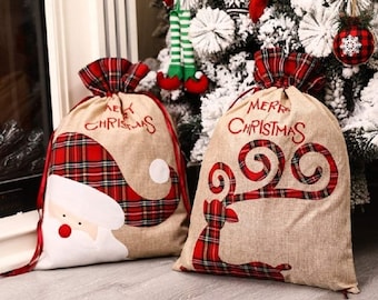 May include: Two Christmas gift bags made of burlap with red and black plaid accents. One bag features a Santa Claus design and the other features a reindeer design. Both bags have the text "Merry Christmas" printed on them.