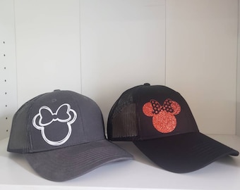 Peut inclure: Deux casquettes de camionneur en maille noire avec un motif Minnie Mouse blanc et rouge. La casquette de gauche a un contour blanc de Minnie Mouse avec un nœud blanc. La casquette de droite a une Minnie Mouse rouge pailletée avec un nœud noir et des pois blancs.