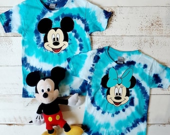 Peut inclure: Deux t-shirts tie-dye représentant Mickey et Minnie Mouse. Les t-shirts sont bleus et blancs avec un motif en spirale. Un jouet en peluche Mickey Mouse est au premier plan.