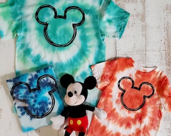 Peut inclure: Quatre t-shirts tie-dye aux tons de bleu, vert et orange, chacun orné d'un contour noir de Mickey Mouse. Une peluche Mickey Mouse est également présente, avec un short rouge, des gants blancs et des oreilles noires. Les chemises sont sur une surface en bois blanc.