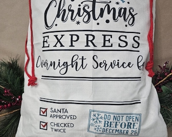 Peut inclure: Sac de Noël en toile blanche avec cordon de serrage rouge, avec le texte "Christmas Express Overnight Service for" et une liste de contrôle avec le texte "Santa Approved Checked Twice". Un tampon bleu avec des flocons de neige indique "Do Not Open Before December 25".
