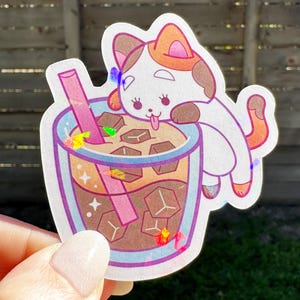 May include: A white cat with orange ears and a pink nose is leaning into a glass of iced coffee with a pink straw. The coffee has ice cubes and is decorated with colorful sprinkles.