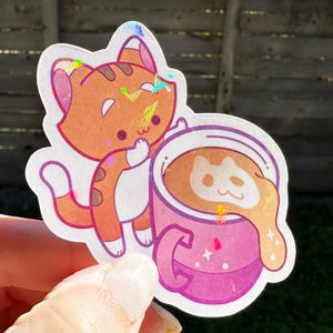 May include: A sticker featuring a cartoon orange cat with a pink coffee mug with a brown cat face design in the foam. The mug has a pink handle and white stars.