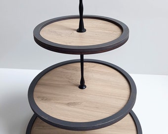 May include: A three-tiered serving stand with a black metal frame and handle. The round tiers have a light wood grain appearance with a dark gray border. The stand is ideal for displaying desserts or appetizers.