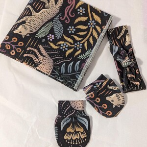 May include: A black and white baby set with a floral pattern featuring wolves. The set includes a blanket, headband, and two mittens.