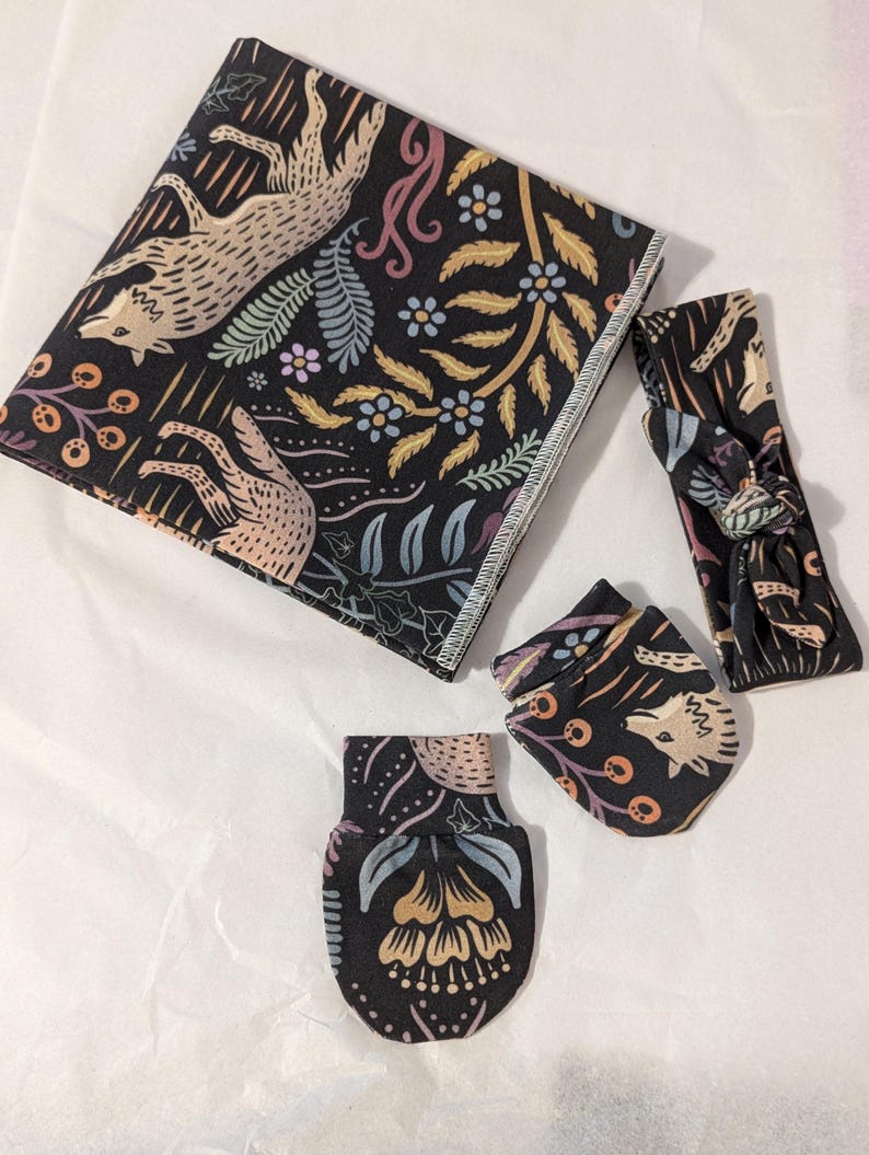 May include: A black and white baby set with a floral pattern featuring wolves. The set includes a blanket, headband, and two mittens.