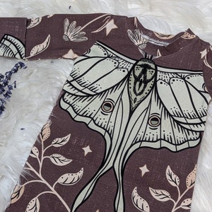 May include: A brown baby sleeper with a large green and white moth design. The moth has a detailed pattern and is surrounded by smaller floral and leaf designs.