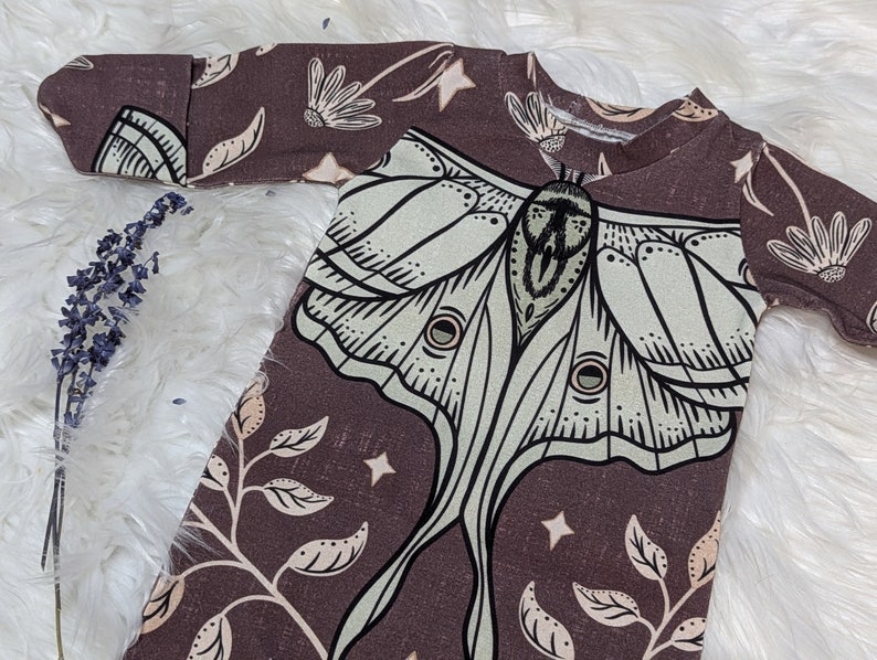 May include: A brown baby sleeper with a large green and white moth design. The moth has a detailed pattern and is surrounded by smaller floral and leaf designs.
