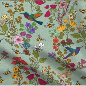 Op de afbeelding: Een tealblauwe stof met een herhalend patroon van kolibries, bijen en bloemen. De kolibries zijn groen, paars en oranje. De bijen zijn geel en zwart. De bloemen zijn roze, rood, geel en wit.