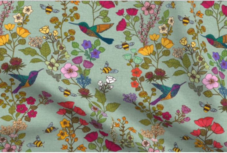 Op de afbeelding: Een tealblauwe stof met een herhalend patroon van kolibries, bijen en bloemen. De kolibries zijn groen, paars en oranje. De bijen zijn geel en zwart. De bloemen zijn roze, rood, geel en wit.