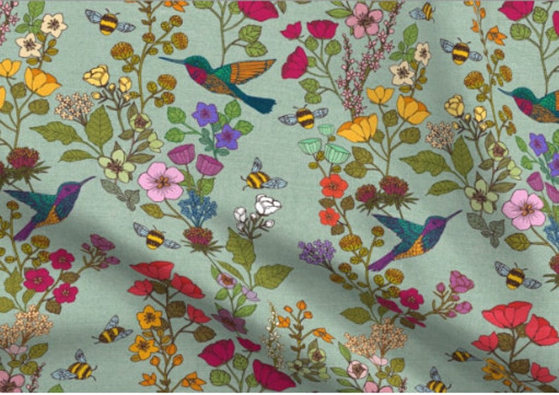 Op de afbeelding: Een tealblauwe stof met een herhalend patroon van kolibries, bijen en bloemen in verschillende kleuren, waaronder roze, geel, rood en groen.