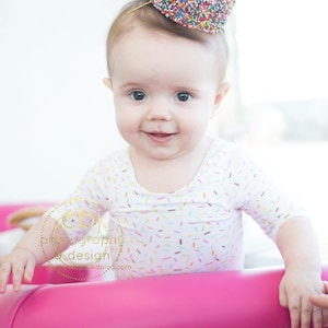 May include: A baby wearing a white and colorful sprinkle-patterned onesie and a colorful sprinkle crown sits on a pink chair.