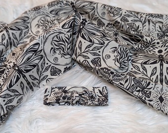 May include: A black and white patterned headband with a dragonfly and moon design. The headband is made of fabric and is tied around the head.