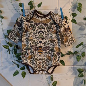May include: A long-sleeved baby onesie with a cream background and a whimsical pattern of mushrooms, bees, butterflies, and floral elements in shades of brown, green, yellow, and pink.
