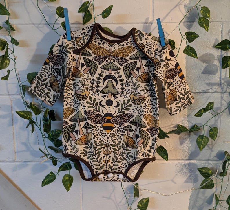 May include: A long-sleeved baby onesie with a cream background and a whimsical pattern of mushrooms, bees, butterflies, and floral elements in shades of brown, green, yellow, and pink.