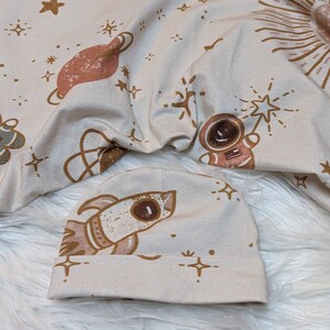 Puede incluir: Un gorro de bebé de tela beige con un diseño de cohete marrón y fondo blanco. El gorro tiene una pequeña estrella marrón y un diseño de estrella estrellada marrón.