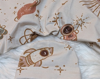 May include: A beige fabric baby hat with a brown rocket ship design and a white background. The hat has a small brown star and a brown starburst design.