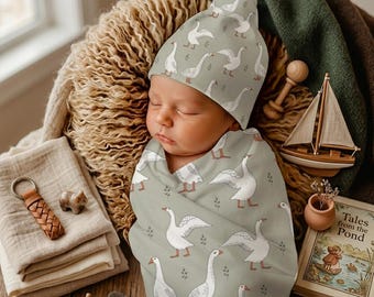 May include: A baby swaddled in a sage green blanket featuring a white goose pattern, wearing a matching beanie. The fabric displays multiple white geese in various poses with small green sprigs. The swaddle and hat set is made of soft fabric.
