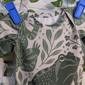 May include: A light pink t-shirt with a green frog and leaf pattern. The frog is stylised and has a detailed design.