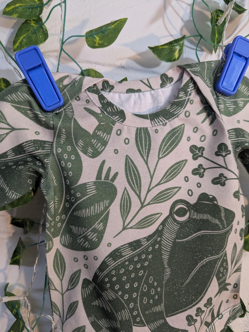 May include: A light pink t-shirt with a green frog and leaf pattern. The frog is stylised and has a detailed design.