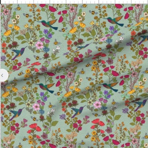 Op de afbeelding: Een tealblauwe stof met een herhalend patroon van kolibries, bijen en kleurrijke bloemen. De kolibries vliegen en de bijen zoemen rond de bloemen.
